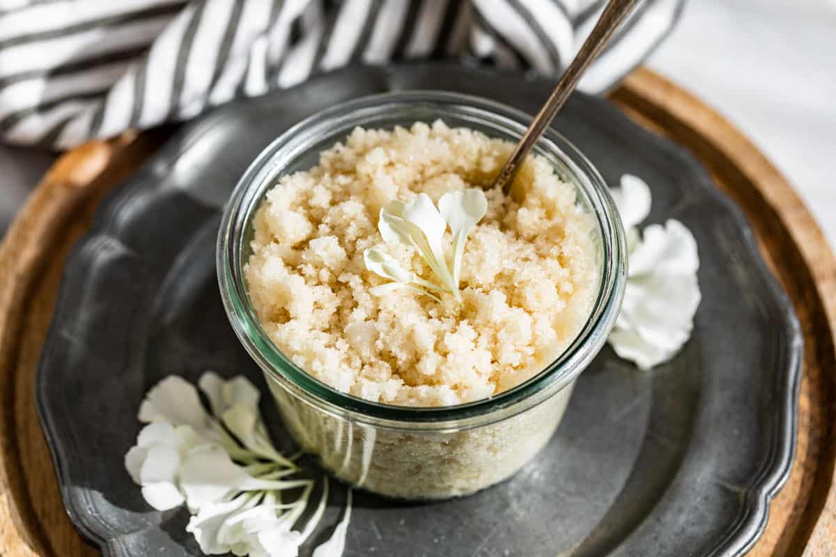 Finished sugar scrub in a glass container with flower petals on top placed on a silver plate on a wooden board.