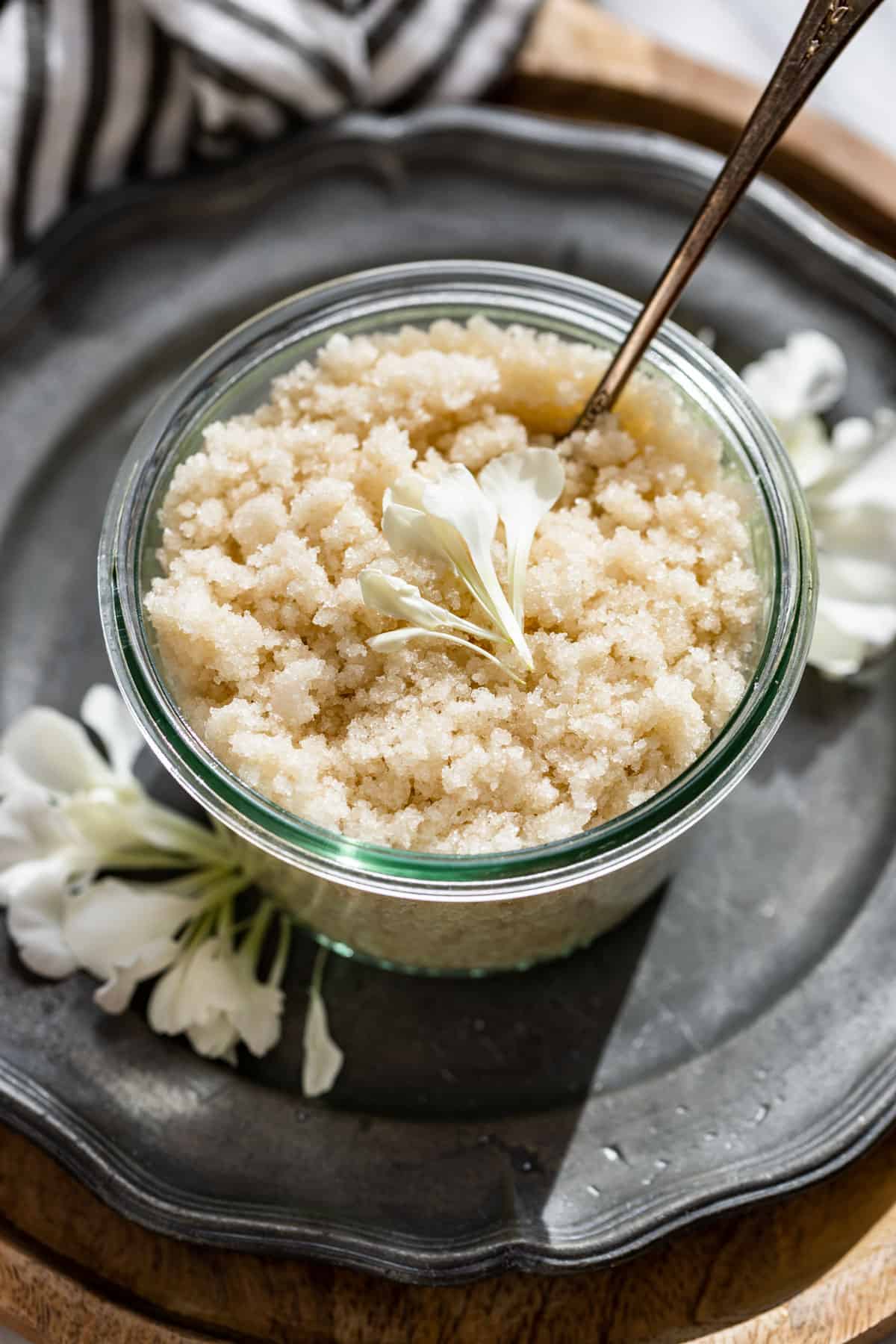Homemade Sugar Scrub in a glass container with flower petals around it.