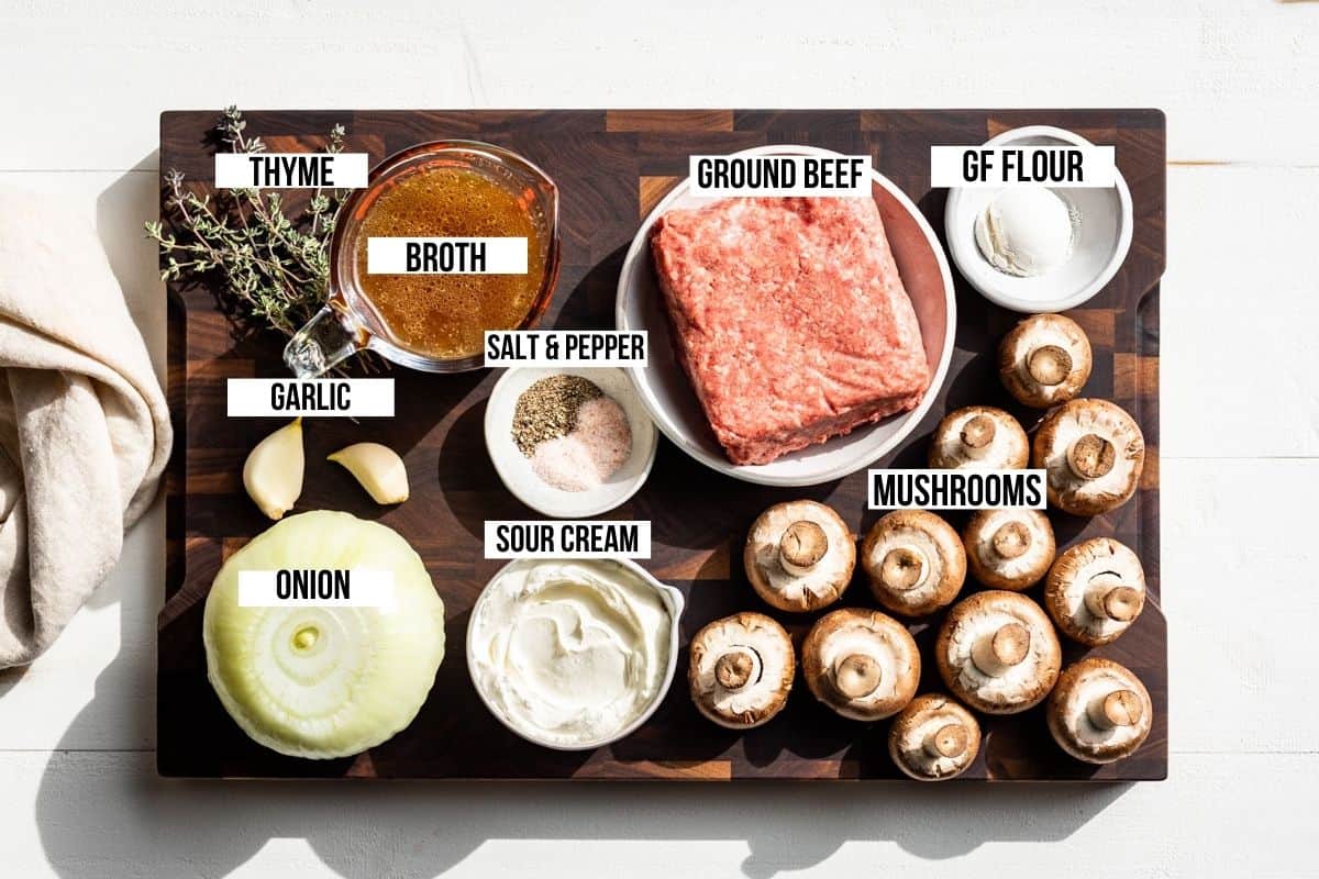 Ground beef, onion, garlic, thyme, beef broth, mushrooms, and sour cream on a wooden cutting board.