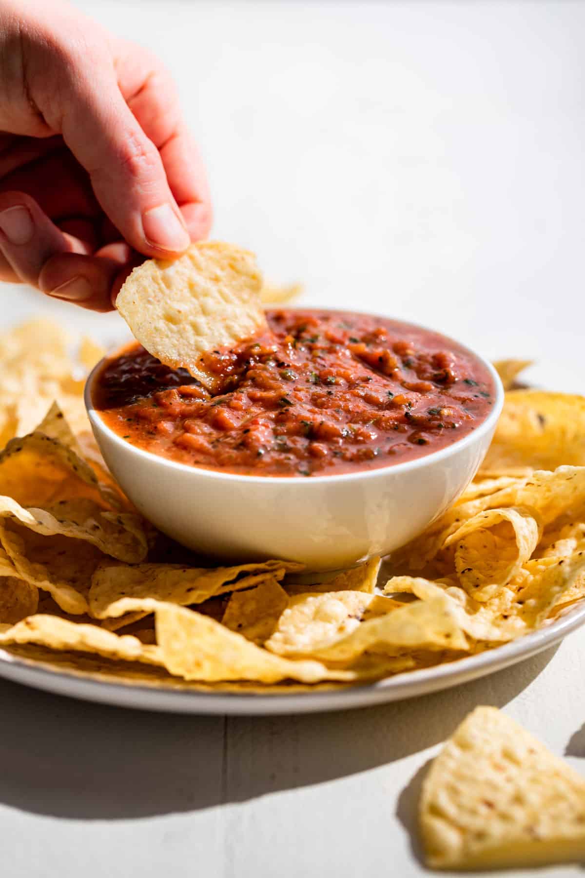 A hand holding a corn chip dipping salsa out of a white bowl with corn chips around it.