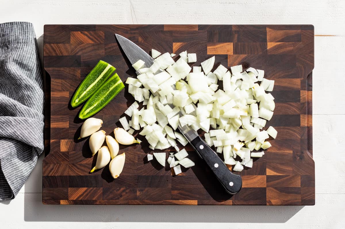 Chopped onion, garlic cloves, and a halved jalapeno on a wood cutting board.
