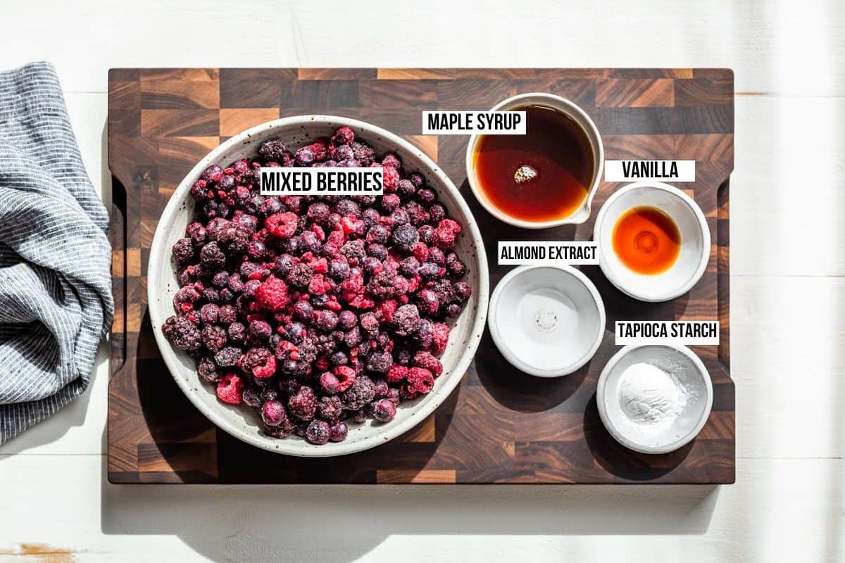 Frozen berries, maple syrup, vanilla extract, almond extract, and tapioca starch in bowls on a wooden cutting board.
