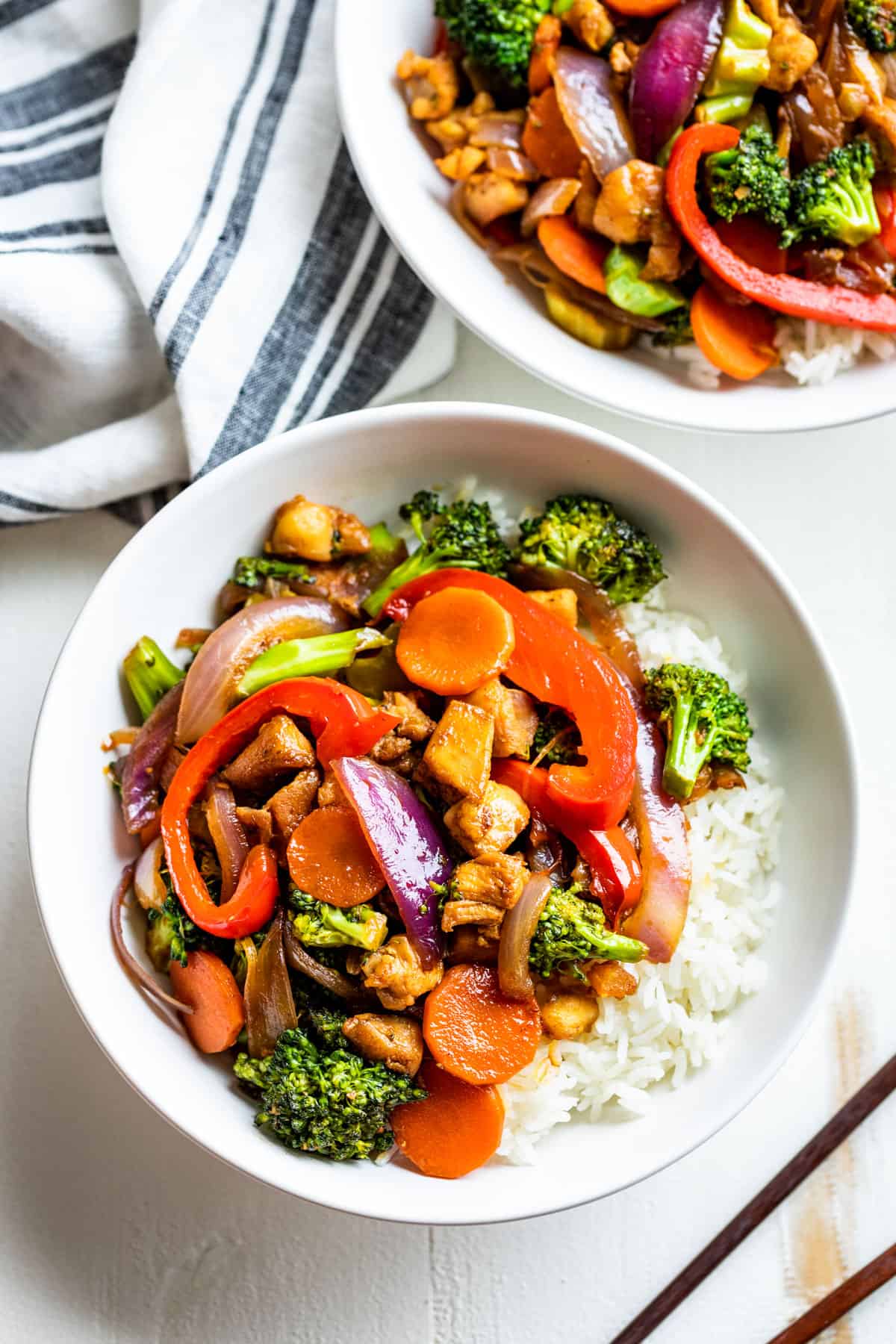 Straight down view of Chicken Stir Fry in white bowls with rice and a blue and white striped linen on the side.