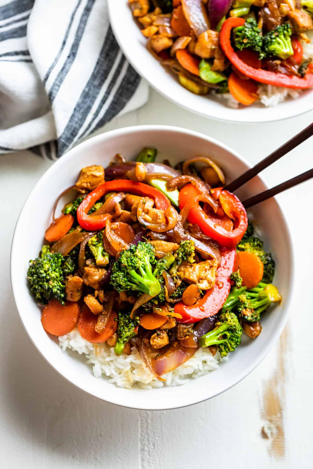 Two white bowls filled with rice and topped with Chicken Stir Fry and a pair of chopsticks.