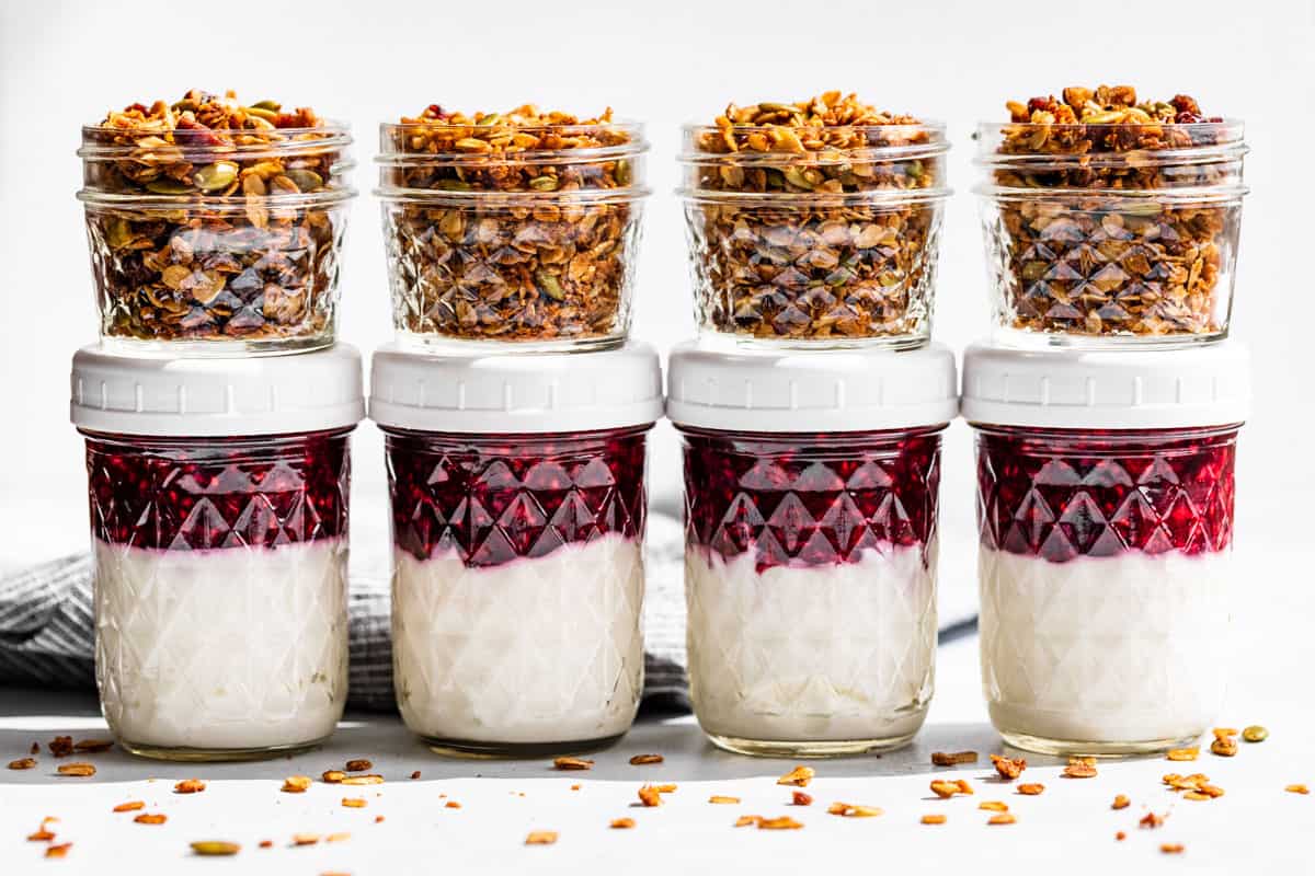 Four glass jars filled halfway with yogurt, topped with fruit sauce and small jars of granola on top.