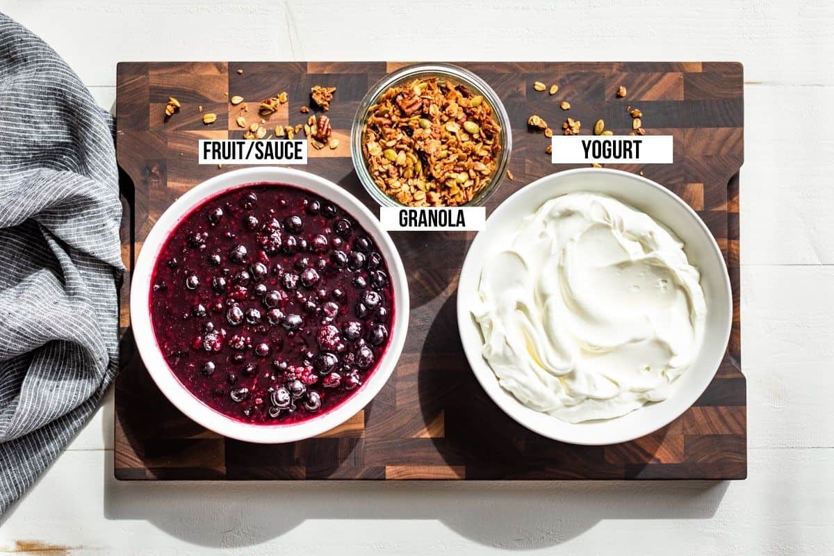 Yogurt, berry sauce, and granola in bowls on a wood cutting board.