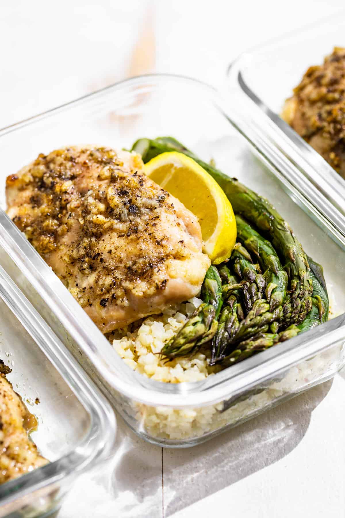 Side view of Garlic Chicken with Asparagus over cauliflower rice topped with a lemon wedge in a glass container.