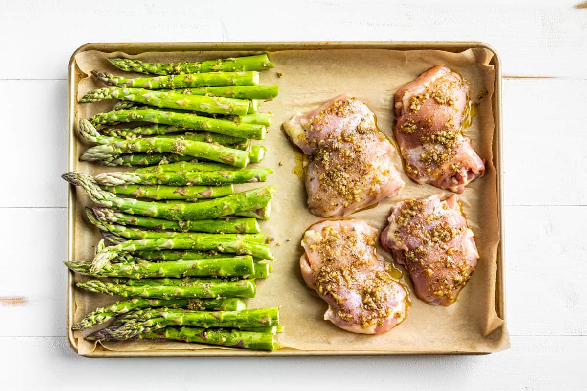 Chicken thighs and asparagus on a parchment lined baking sheet with a garlic olive oil mixture on top.