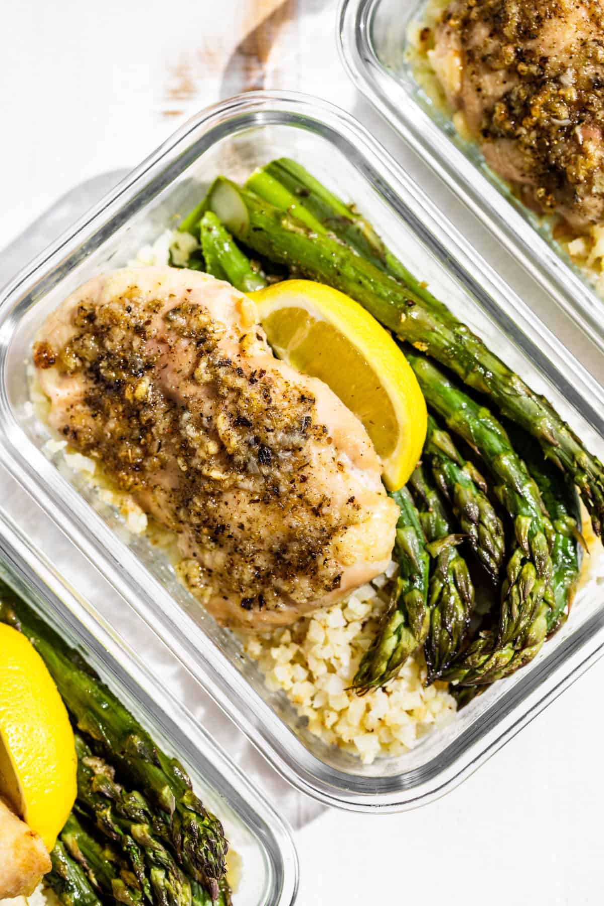 Three clear glass containers with Garlic Herb Chicken, Asparagus, and Cauliflower rice topped with lemon wedges.