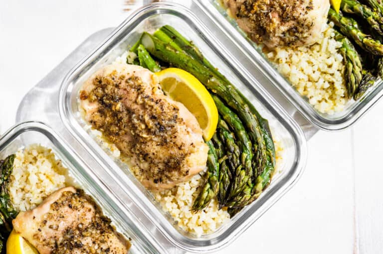 Three glass containers with Garlic Herb Chicken and Asparagus topped with lemon wedges on a white background.