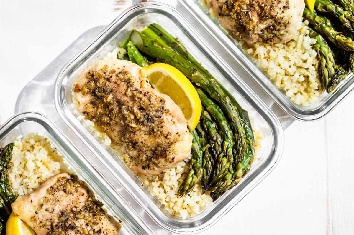 Three glass containers with Garlic Herb Chicken and Asparagus topped with lemon wedges on a white background.