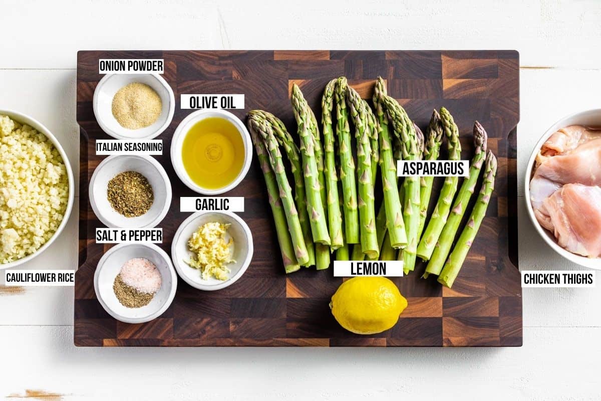 Asparagus, chicken thighs, a lemon, olive oil, spices, garlic, and cauliflower rice in bowls on a wooden cutting board.