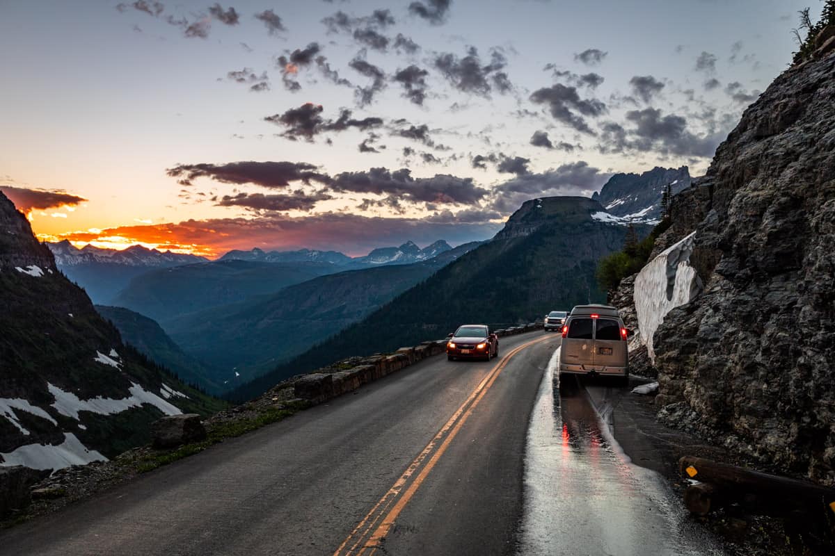 Vehicles on Going to the Sun Road with a mountain and sunset in the background.