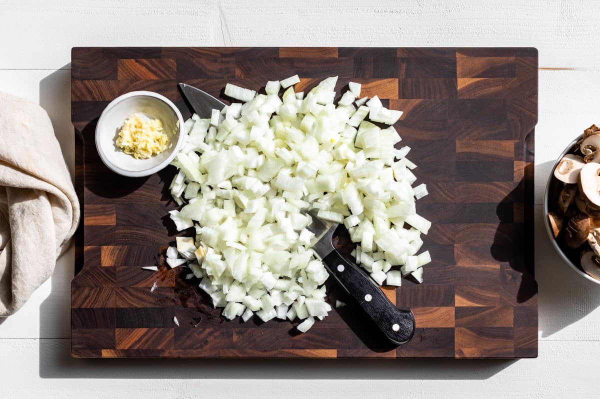 Diced onion, minced garlic, and sliced mushrooms on a wooden cutting board.