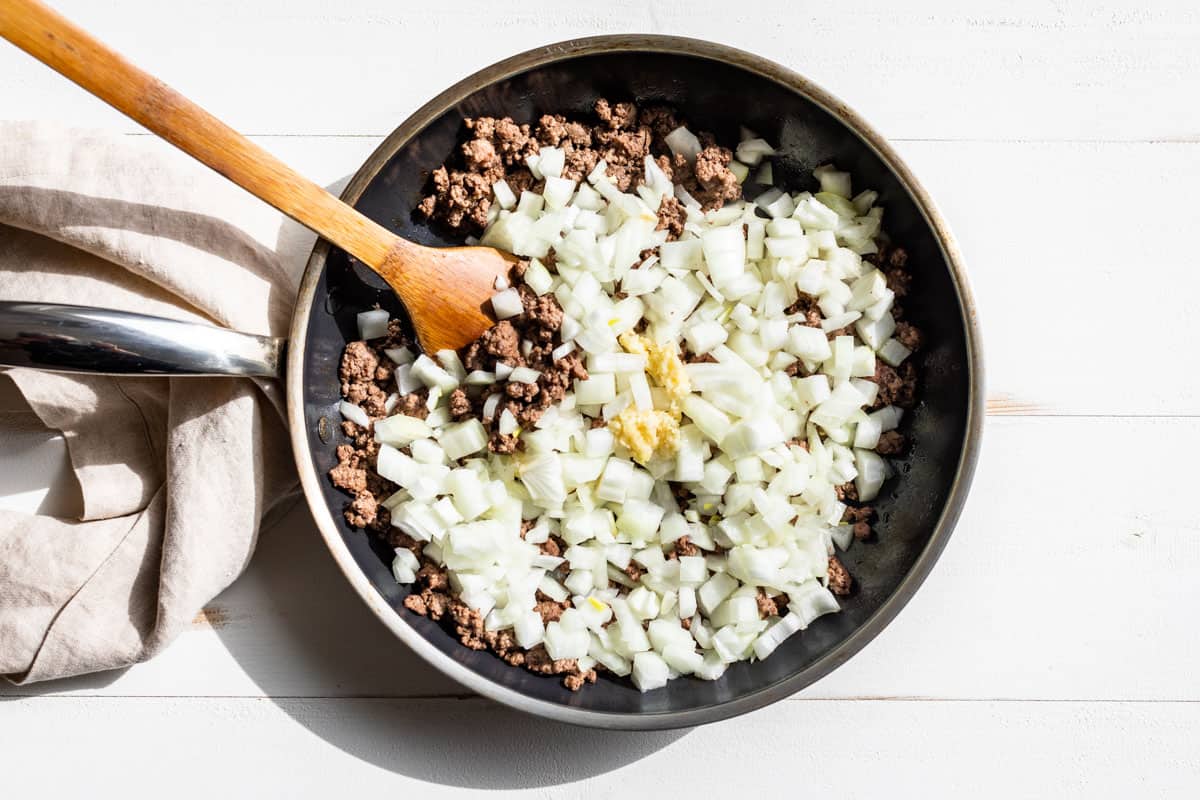 Sautéed ground beef in a skillet with diced onion and garlic being added to the pan.