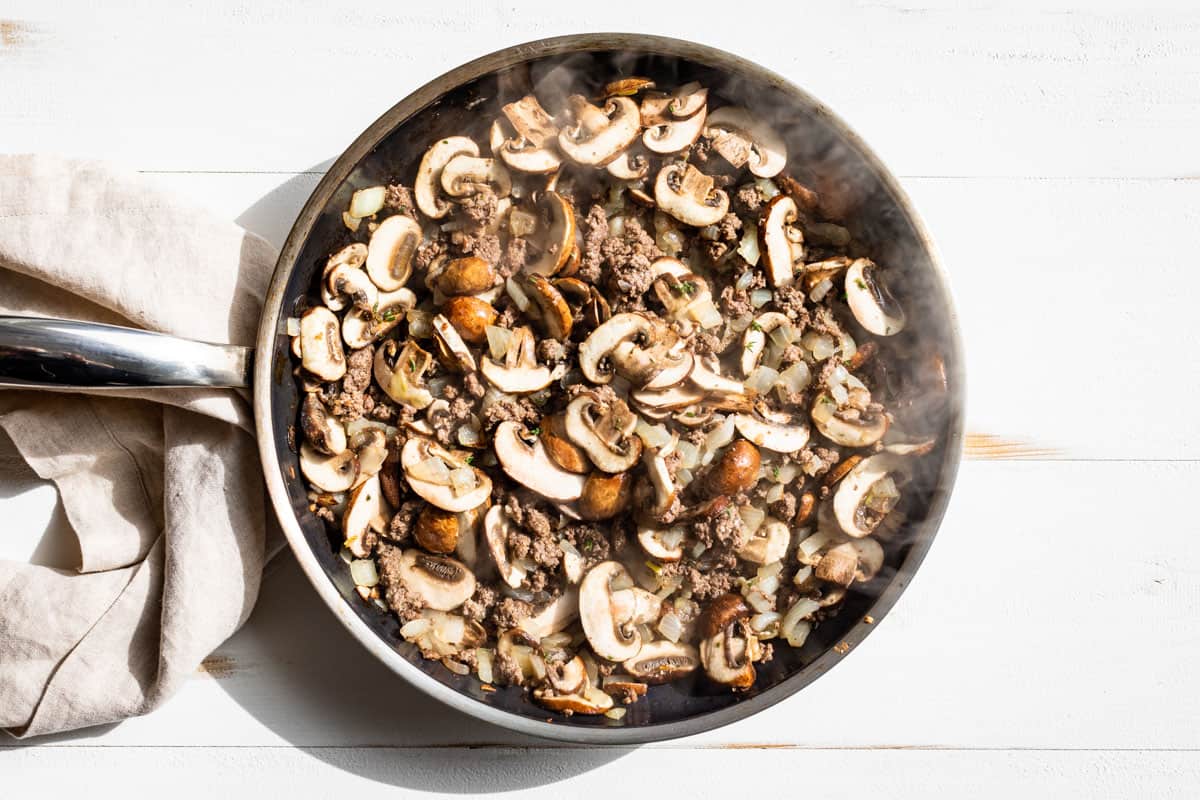 Adding the sliced mushrooms to the sautéed ground beef in a skillet.