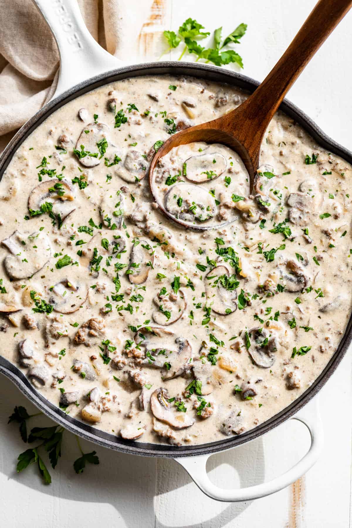Straight down view of Ground Beef Stroganoff in a white skillet with a wooden ladle all sprinkled with parsley.