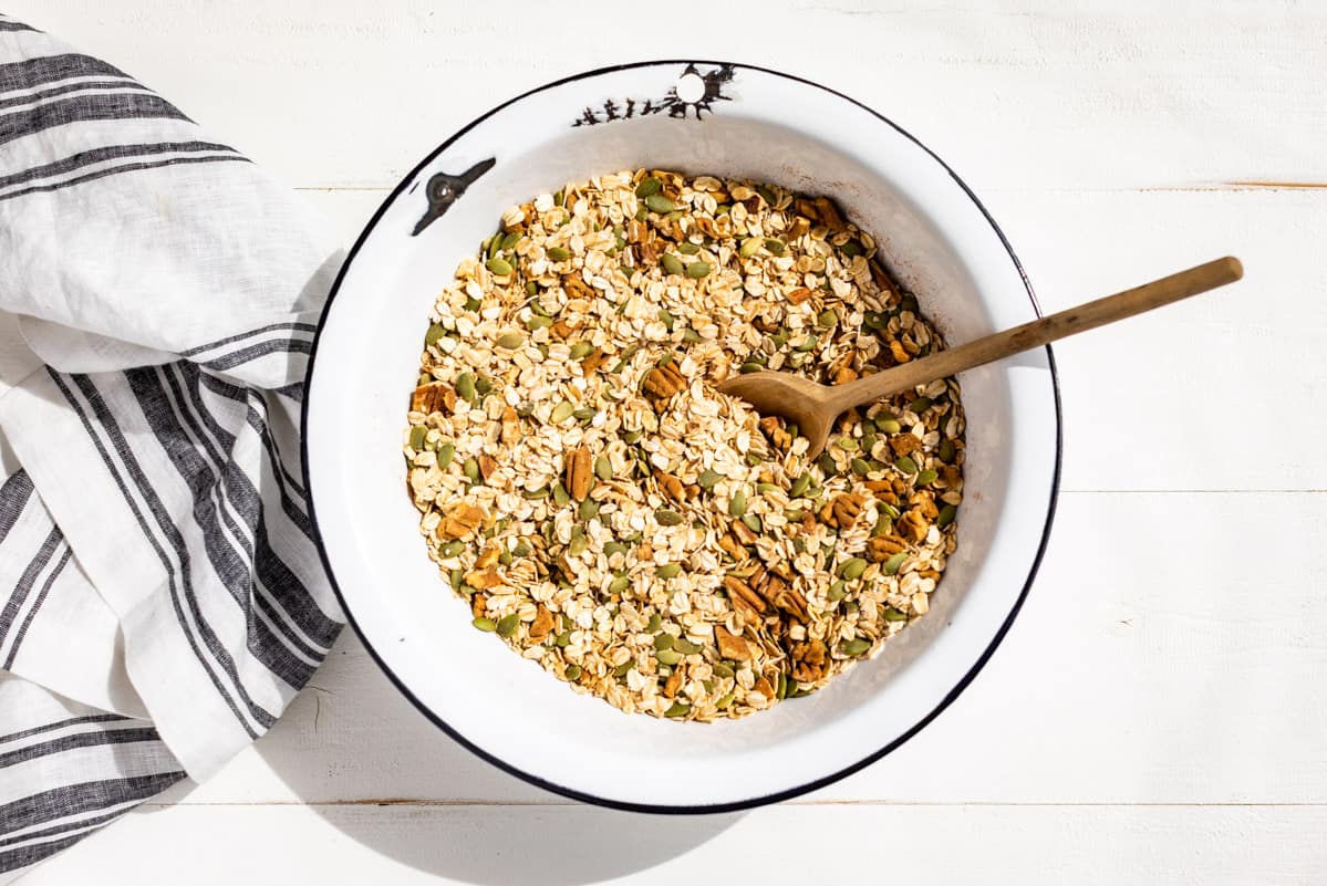 A white enameled mixing bowl with oats, coconut, pecans and pumpkin seeds being mixed together.