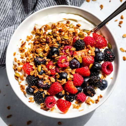 A white bowl with yogurt and berries topped with Homemade Granola and drizzled with honey on a blue background.