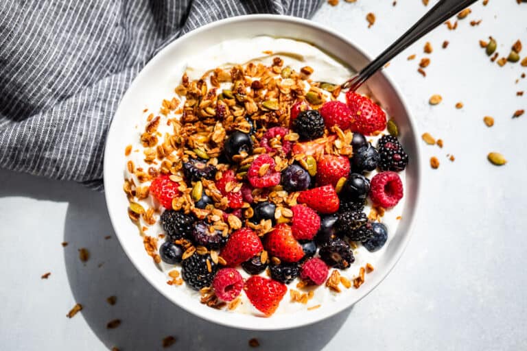 A white bowl with yogurt and berries topped with Homemade Granola and drizzled with honey on a blue background.