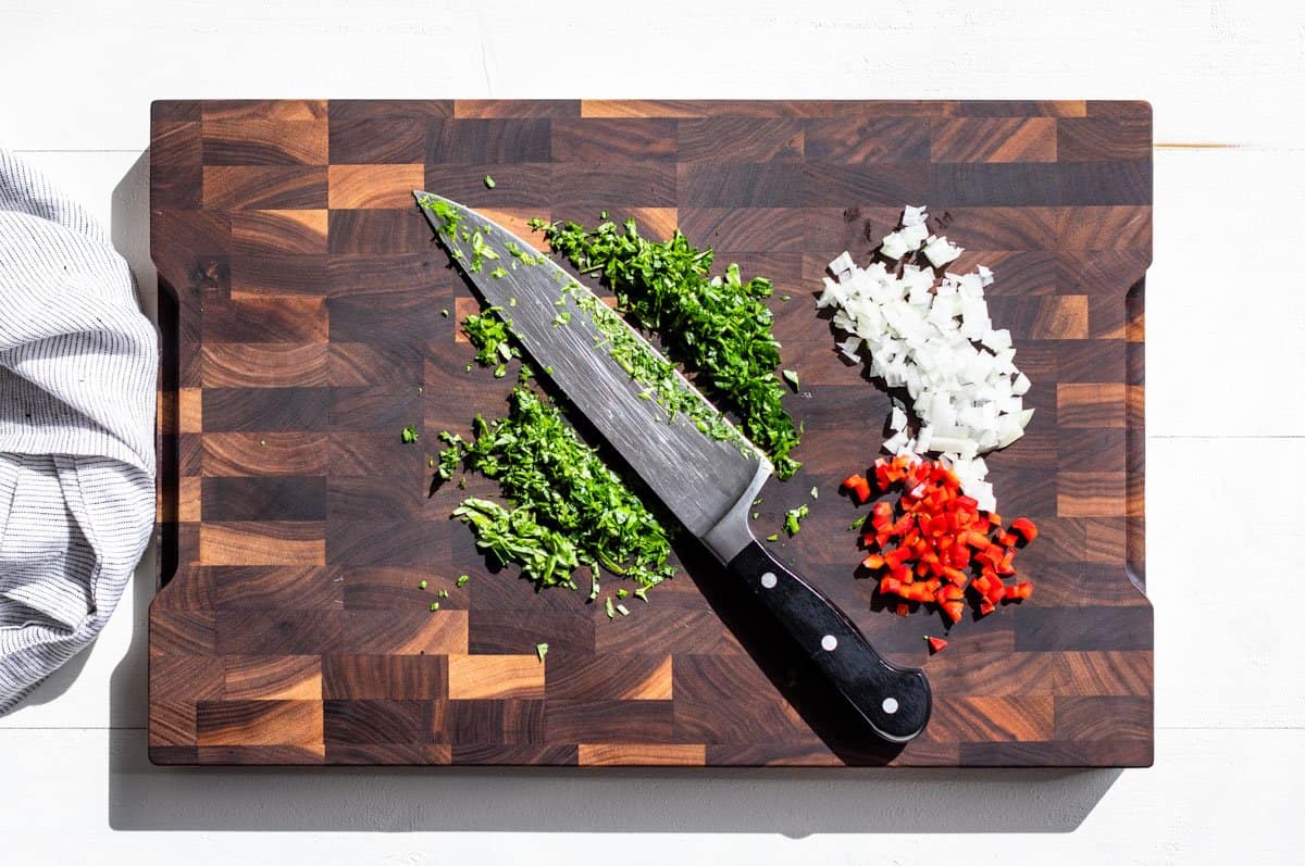 Minced red bell pepper, minced onion, and chopped basil & parsley on a wood cutting board.