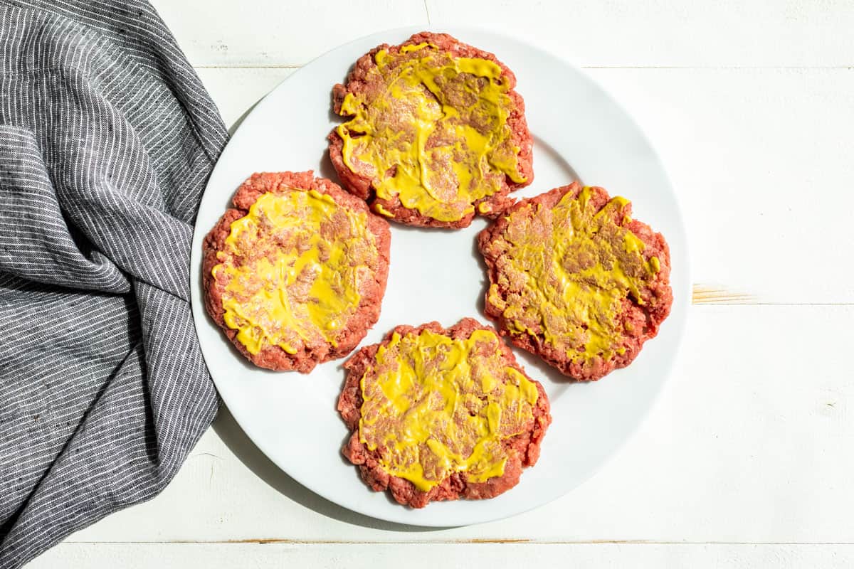 Formed burger patties lightly coated with mustard on 1 side on a white plate.