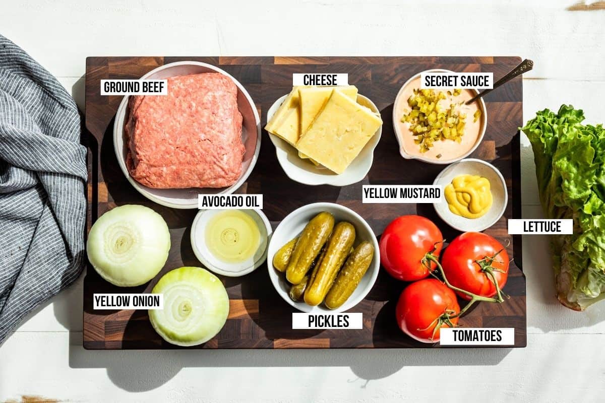 Ground beef, cheddar cheese, onions, pickles, tomatoes, mustard, lettuce, and secret sauce in bowls on a wooden cutting board.