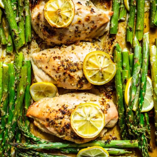 Downwards view of three chicken breasts topped with lemon slices with asparagus being lifted out with a wooden spatula.
