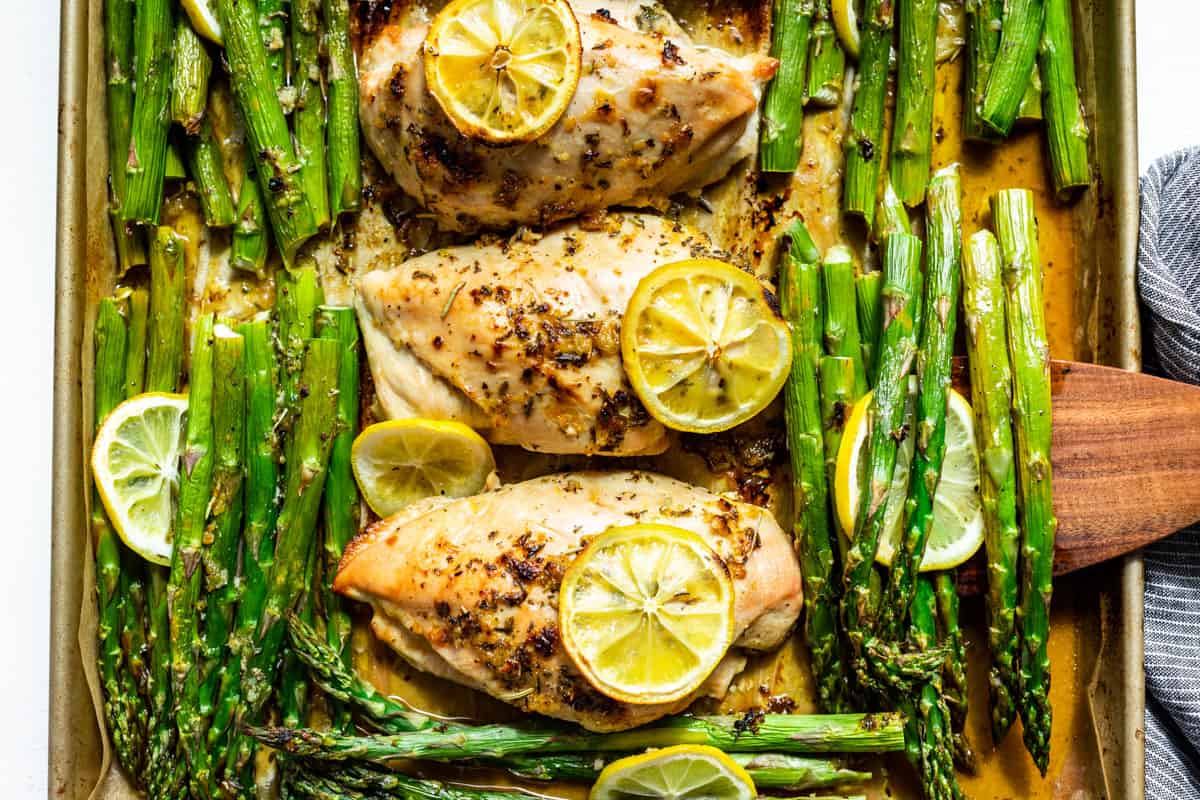 Downwards view of three chicken breasts topped with lemon slices with asparagus being lifted out with a wooden spatula.