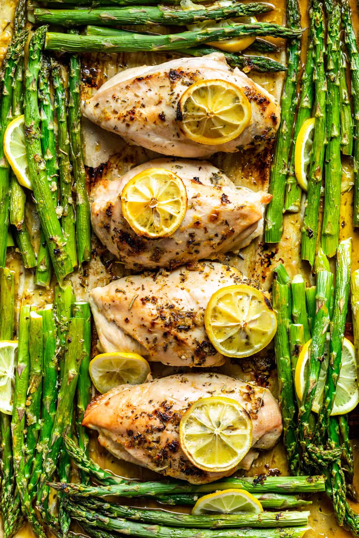 Straight down view of four chicken breasts with asparagus around them and lemon slices on top.