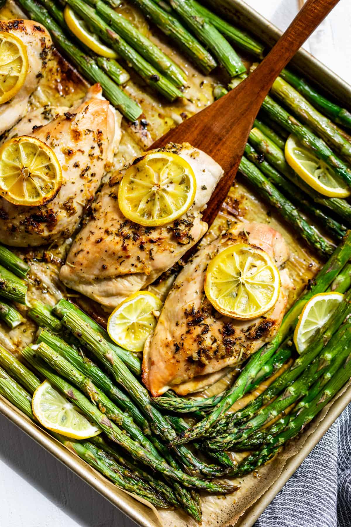 45 degree angle shot of finished Lemon Chicken and Asparagus on a sheet pan with a wooden spatula.