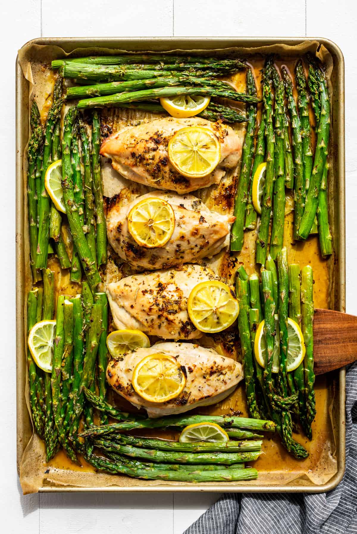 Further out view of Lemon Chicken and Asparagus on a sheet pan with a wooden spatular scooping out some asparagus.