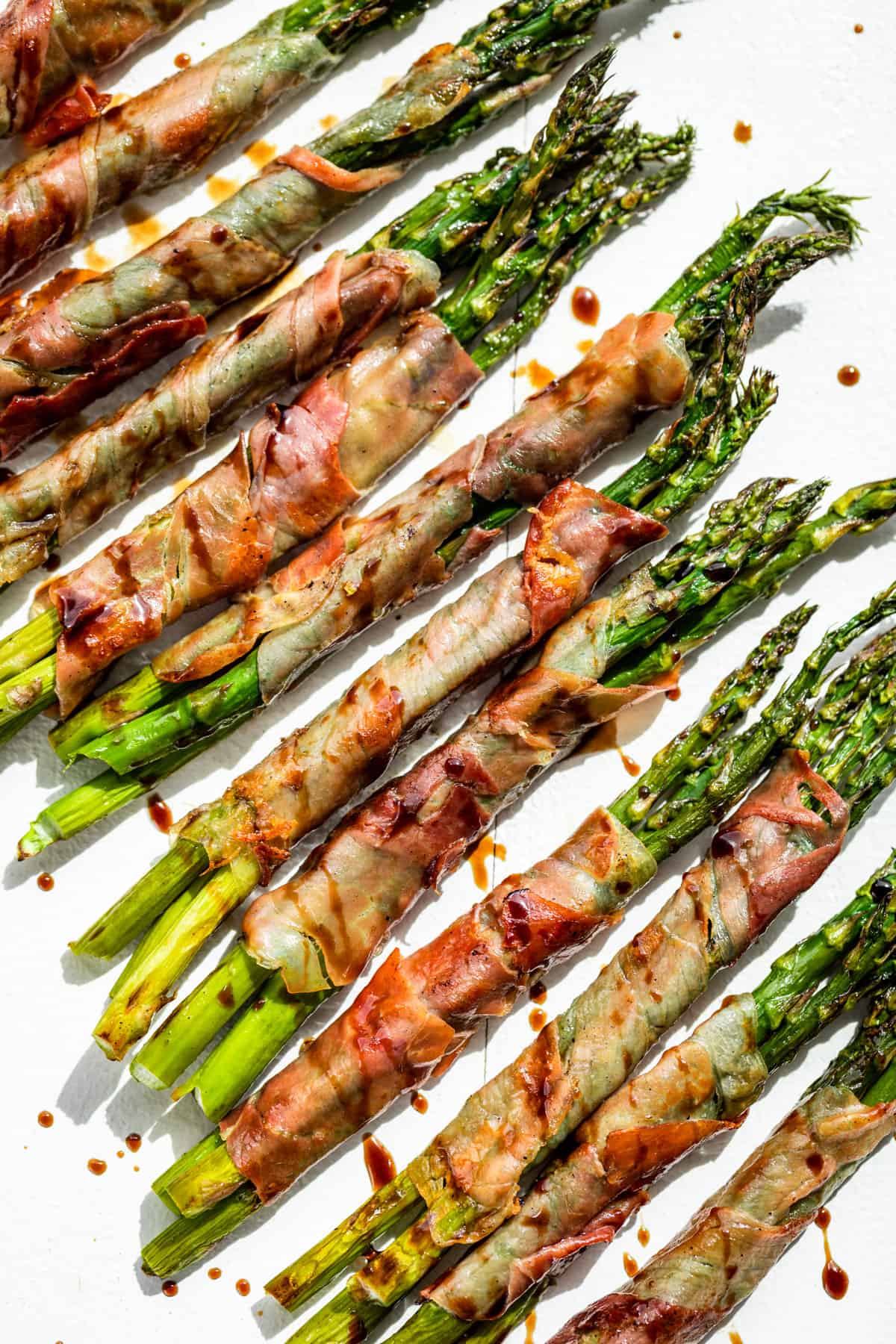 Looking down at Prosciutto Wrapped Asparagus drizzled with reduced balsamic vinegar on a white background.