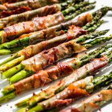 Close view of Prosciutto Wrapped Asparagus on a white painted wood background drizzled with reduced balsamic vinegar.