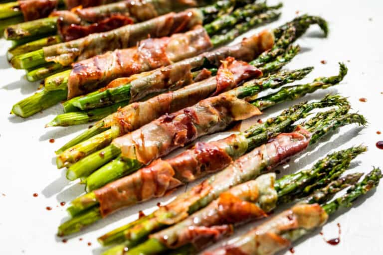 Close view of Prosciutto Wrapped Asparagus on a white painted wood background drizzled with reduced balsamic vinegar.