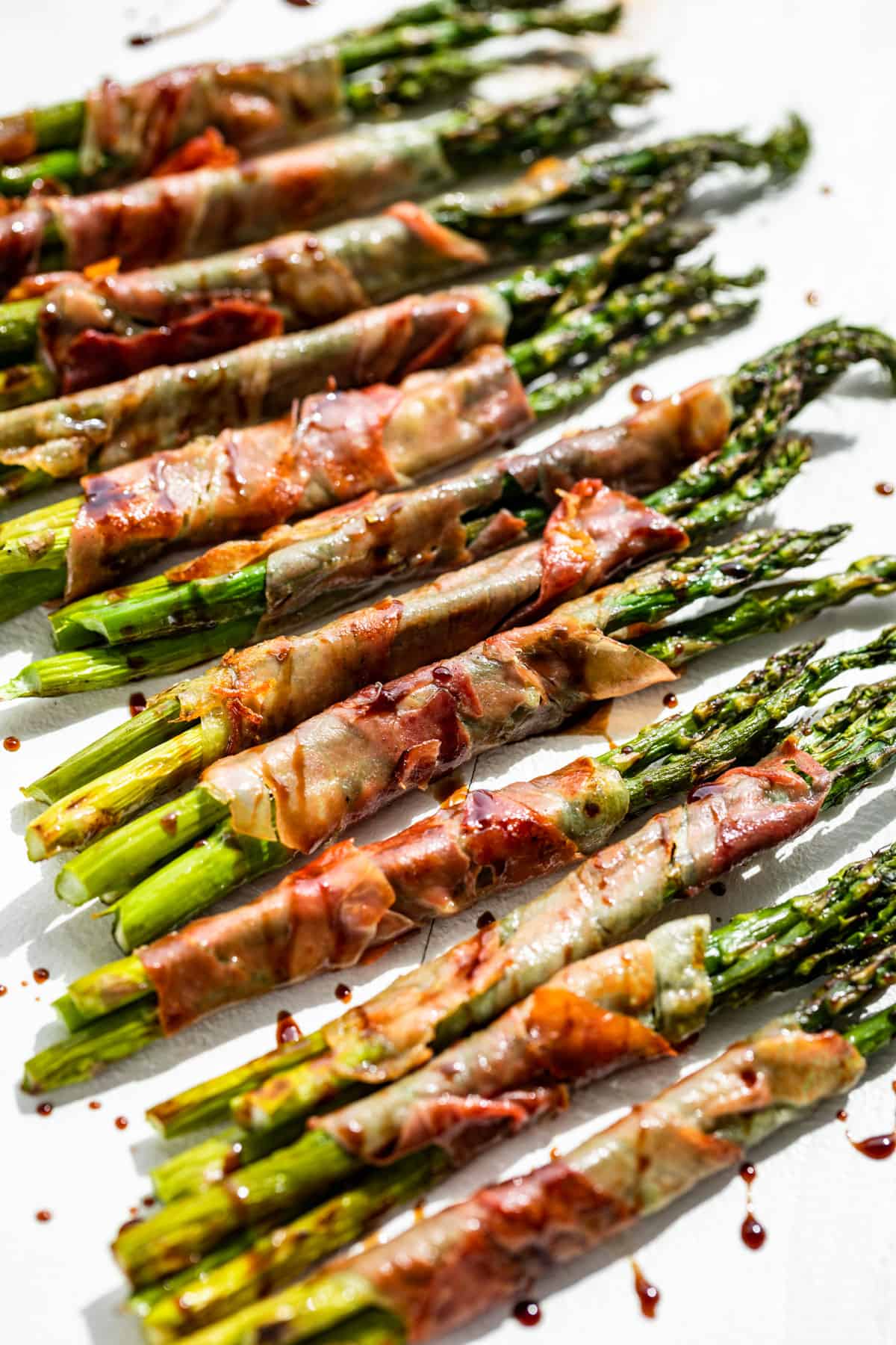 Side view of Prosciutto Wrapped Asparagus drizzled with reduced balsamic vinegar on a white background.