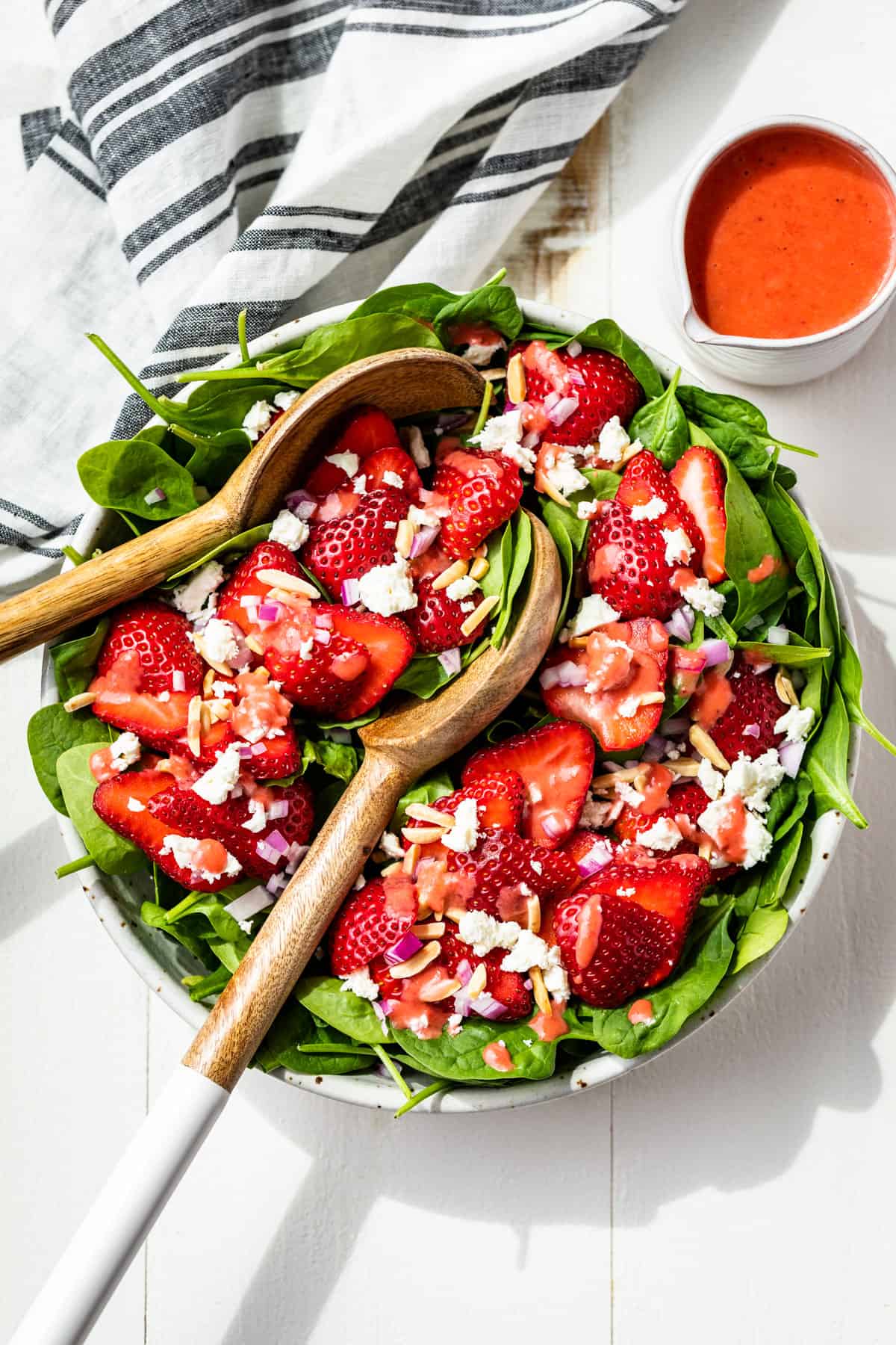 A further out view of the whole Strawberry Spinach Salad in a white bowl with white handled wooden serving spoons, a blue and white striped linen and strawberry vinaigrette on the side.