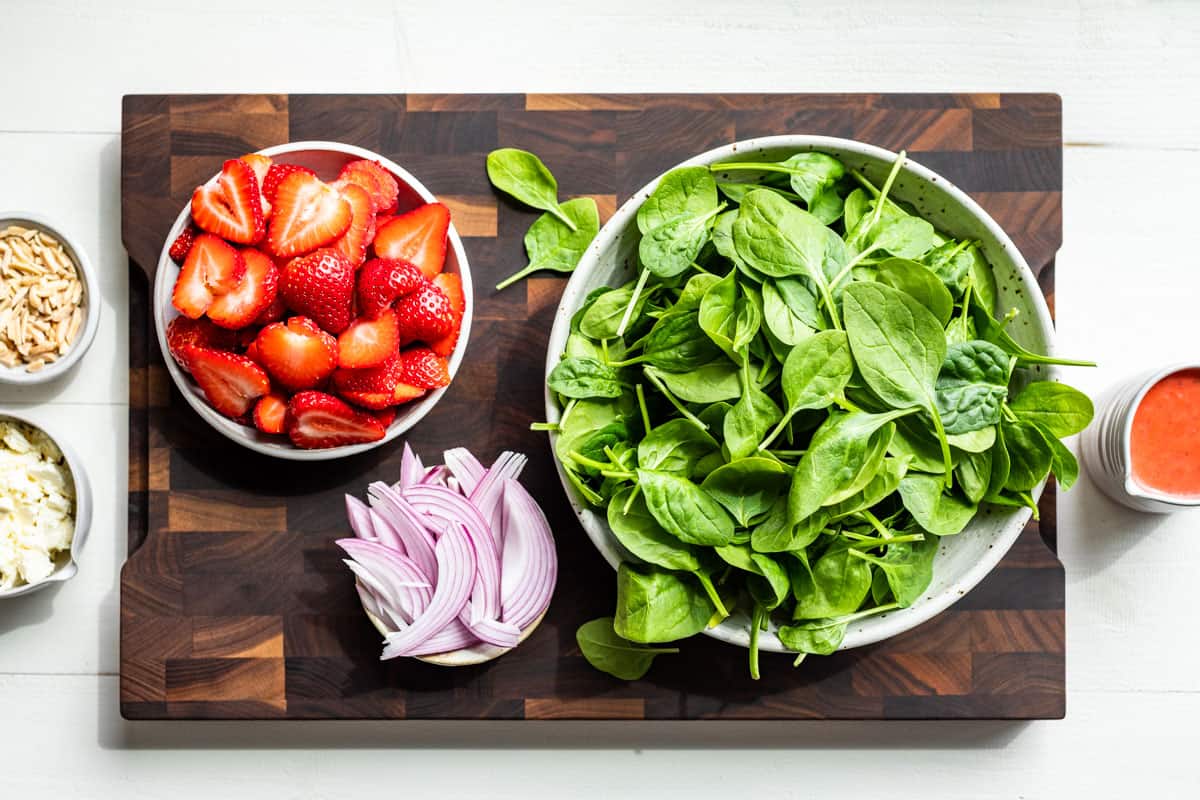 Sliced strawberries, sliced red onion, crumbled feta cheese, spinach, nuts and strawberry vinaigrette in bowls on a wooden cutting board.