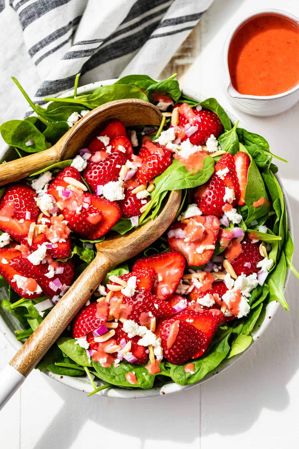 A white bowl filled with Strawberry Spinach Salad with white handled wooden serving spoons and a jar of strawberry vinaigrette.