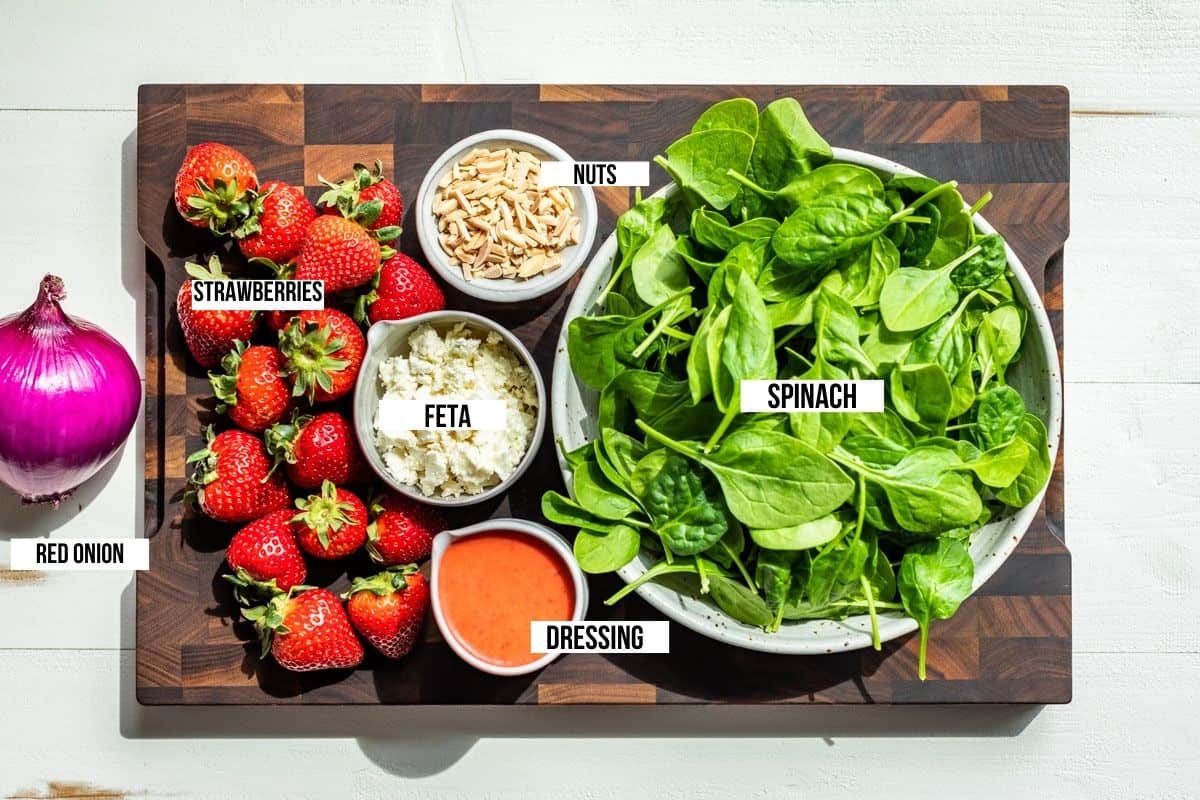 Bowls of spinach, feta cheese, nuts, strawberry vinaigrette, red onion, and strawberries on a wooden cutting board.