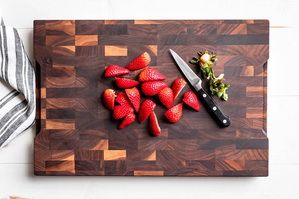 Hulled and quartered strawberries on a wood cutting board with a pairing knife on the side.