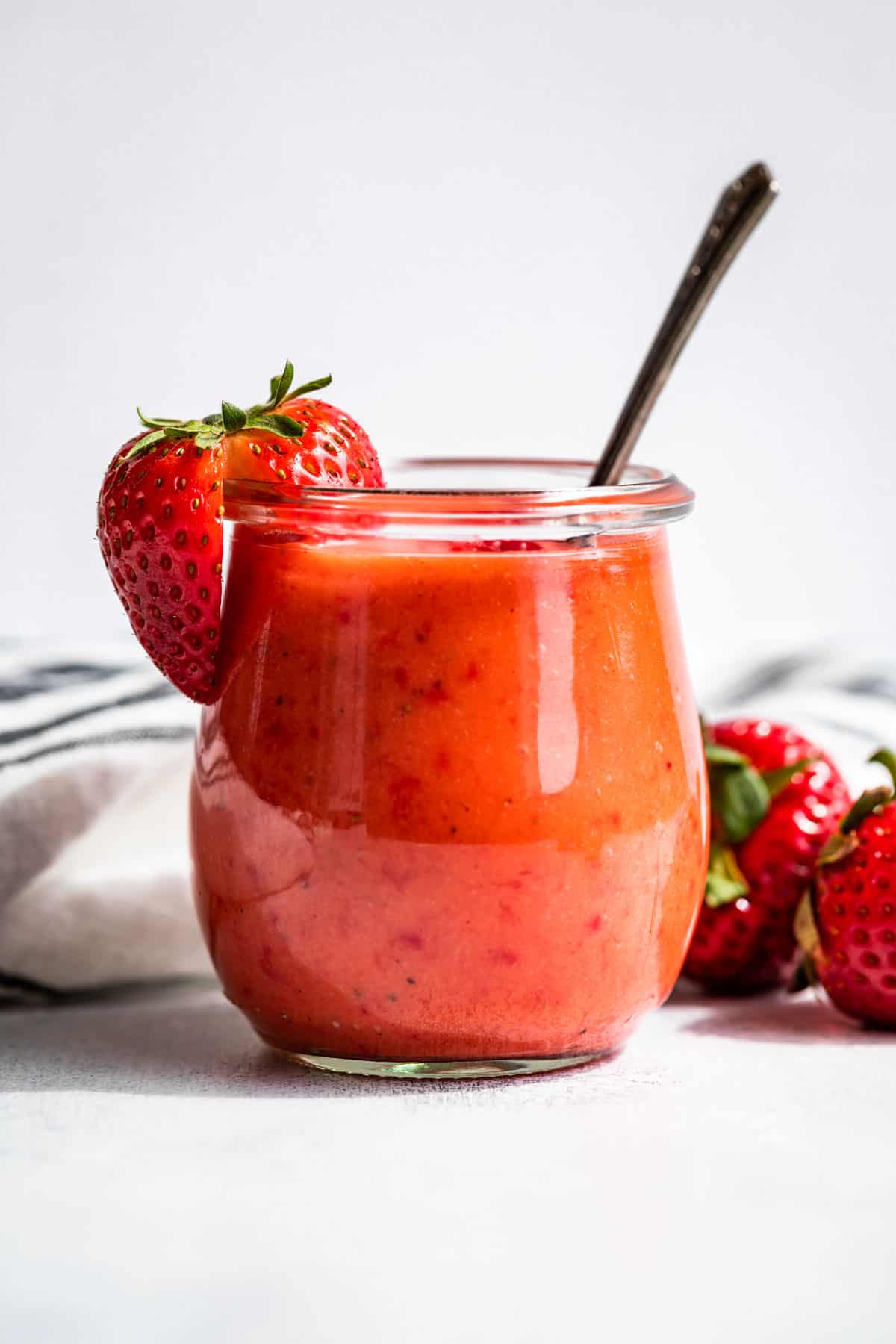 A clear glass jar filled with Strawberry Vinaigrette with a sliced strawberry on the rim.