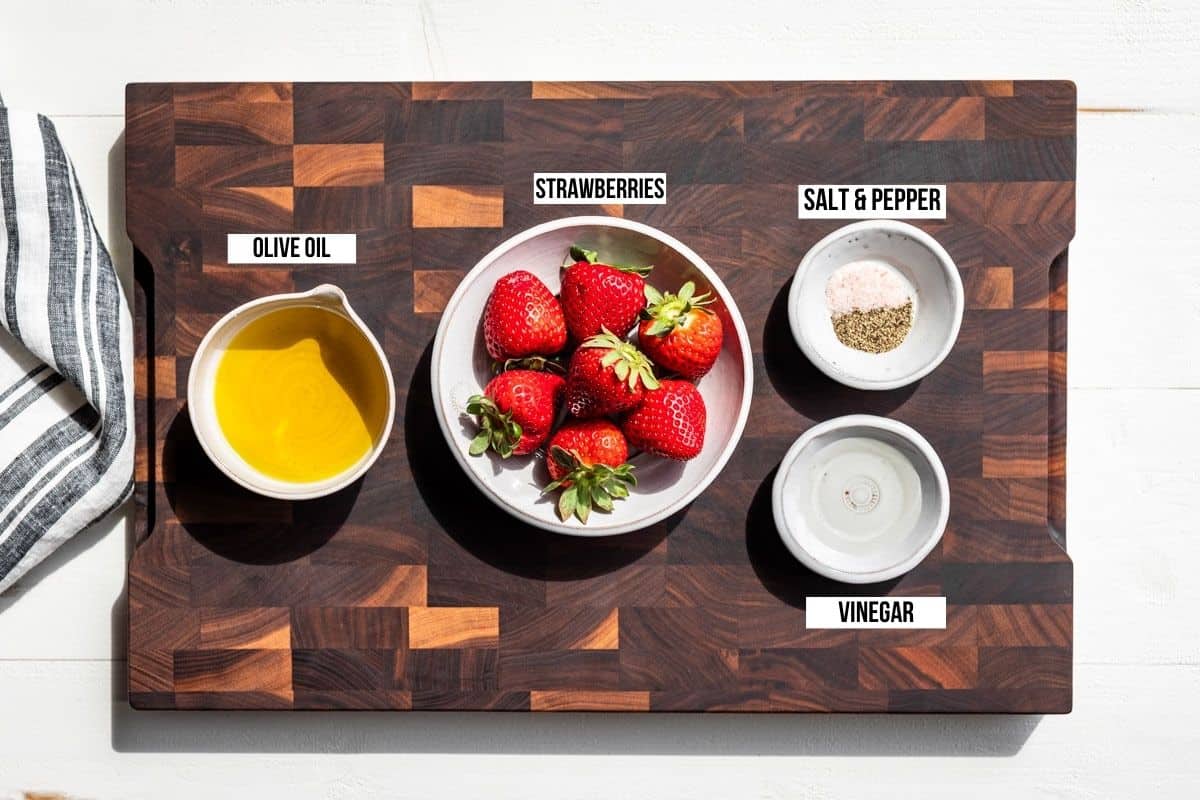 Olive oil, strawberries, sea salt, pepper, and white wine vinegar in small containers on a wood cutting board.