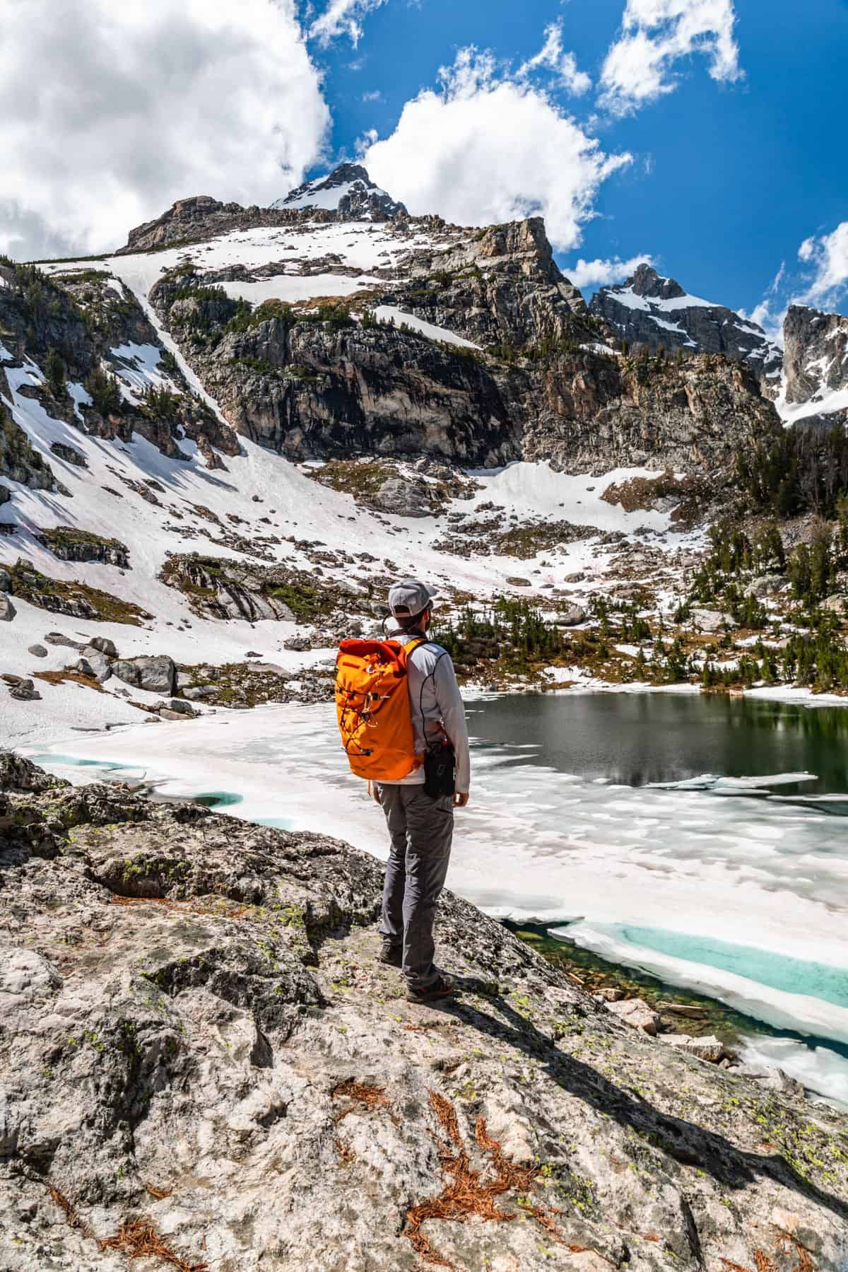 Tall rocky mountain peaks dusted in snow with a partially frozen deep blue lake below and a man with an orange backpack standing on a rock looking at the lake.