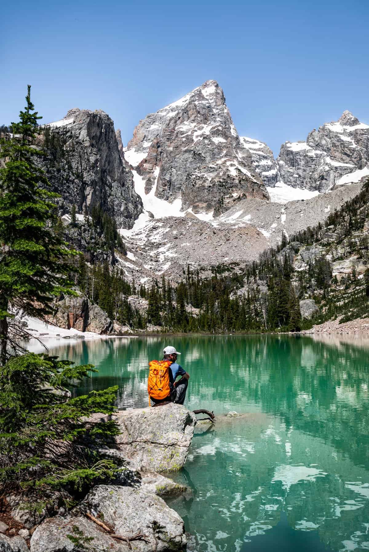 A man with an orange backpack sitting on a large rock looking out over a turquoise lake with a tall mountain peak in the background.