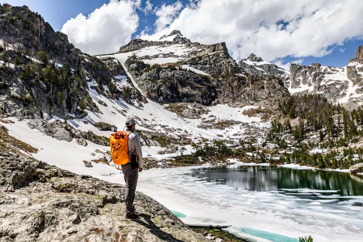 Large mountain cirque with a partially frozen deep blue lake and a man with an orange backpack standing on the rocks looking out.