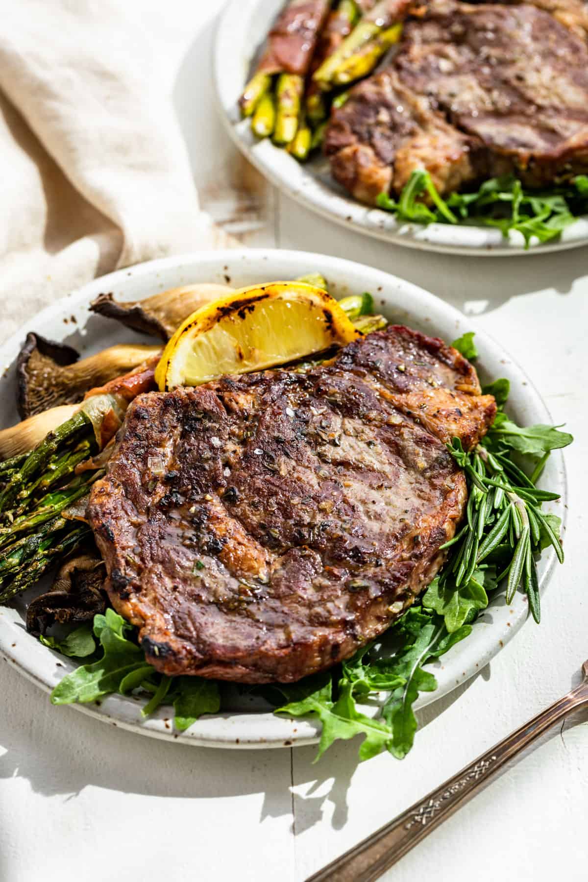 Side view of two plates with Tuscan Steak on a bed of arugula with a lemon wedge, asparagus, and mushrooms on the side.