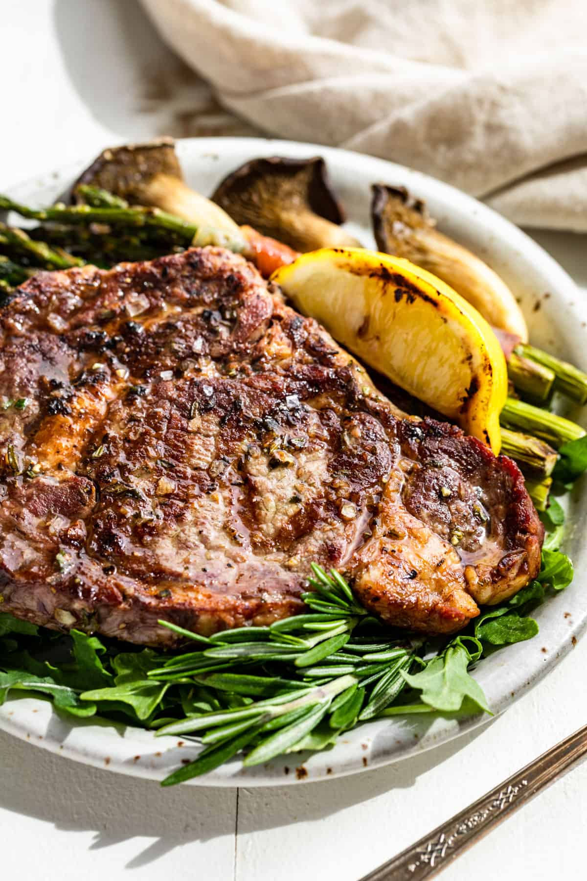 Closer macro side view looking at the finished Tuscan Steak on a bed of arugula with a lemon wedge, asparagus, and mushrooms on the side.