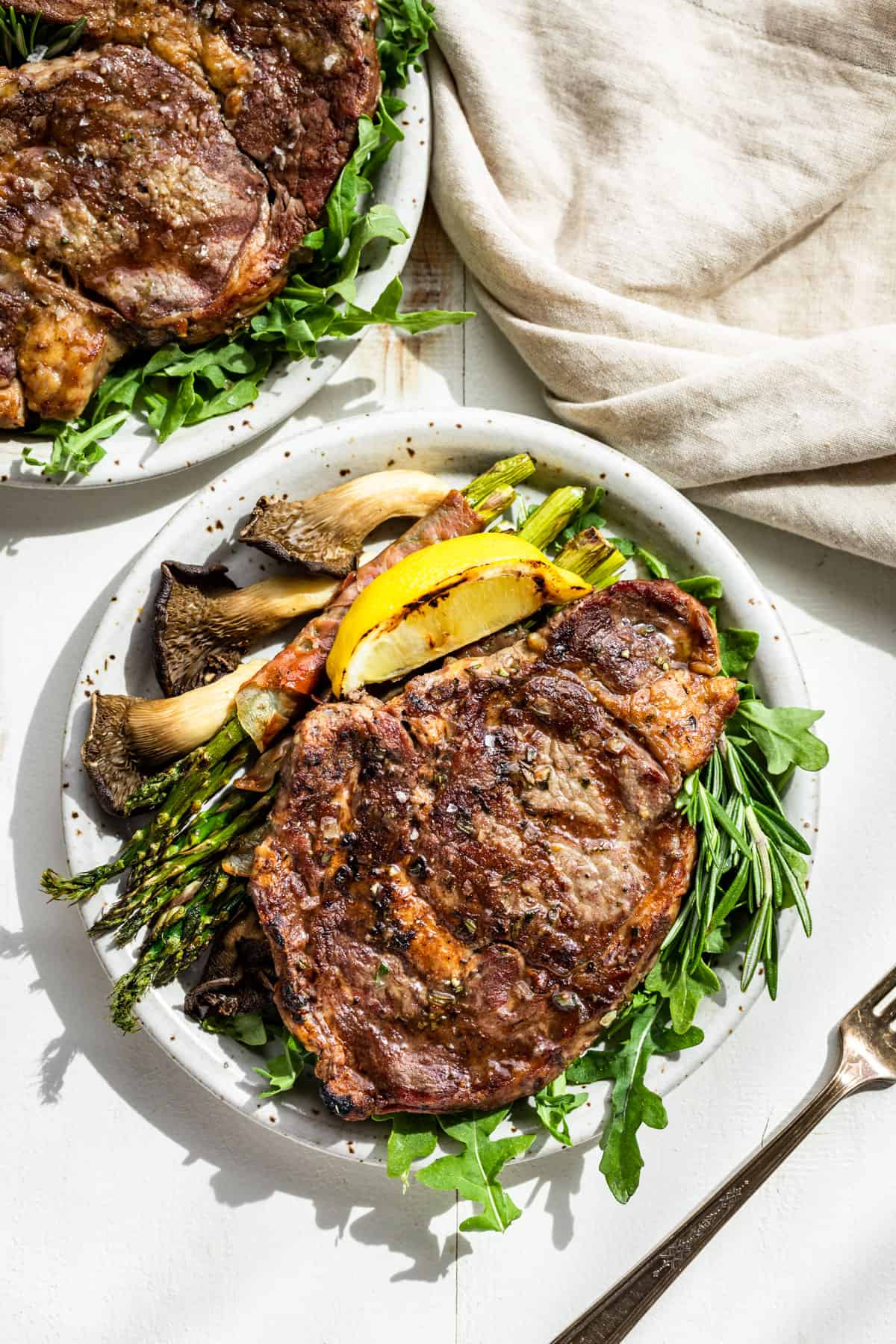 Looking down at two plates with Tuscan Steak on a bed of arugula with a lemon wedge, asparagus, and mushrooms on the side.
