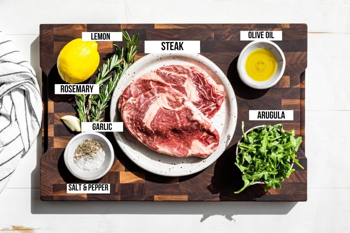 A lemon, two ribeye steaks, olive oil, arugula, sea salt, pepper, and rosemary in containers on a wood cutting board.